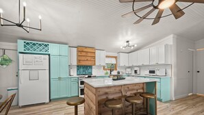 Une cuisine moderne avec des meubles blancs, un îlot central en bois doté de tabourets, un réfrigérateur et un ventilateur de plafond.