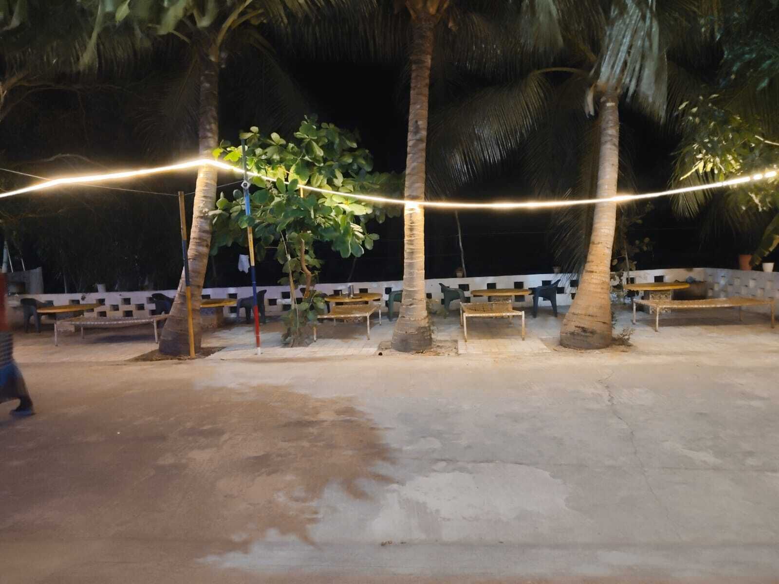 Ein Sitzbereich im Freien mit Bänken und Tischen unter Palmen, beleuchtet von Lichterketten.