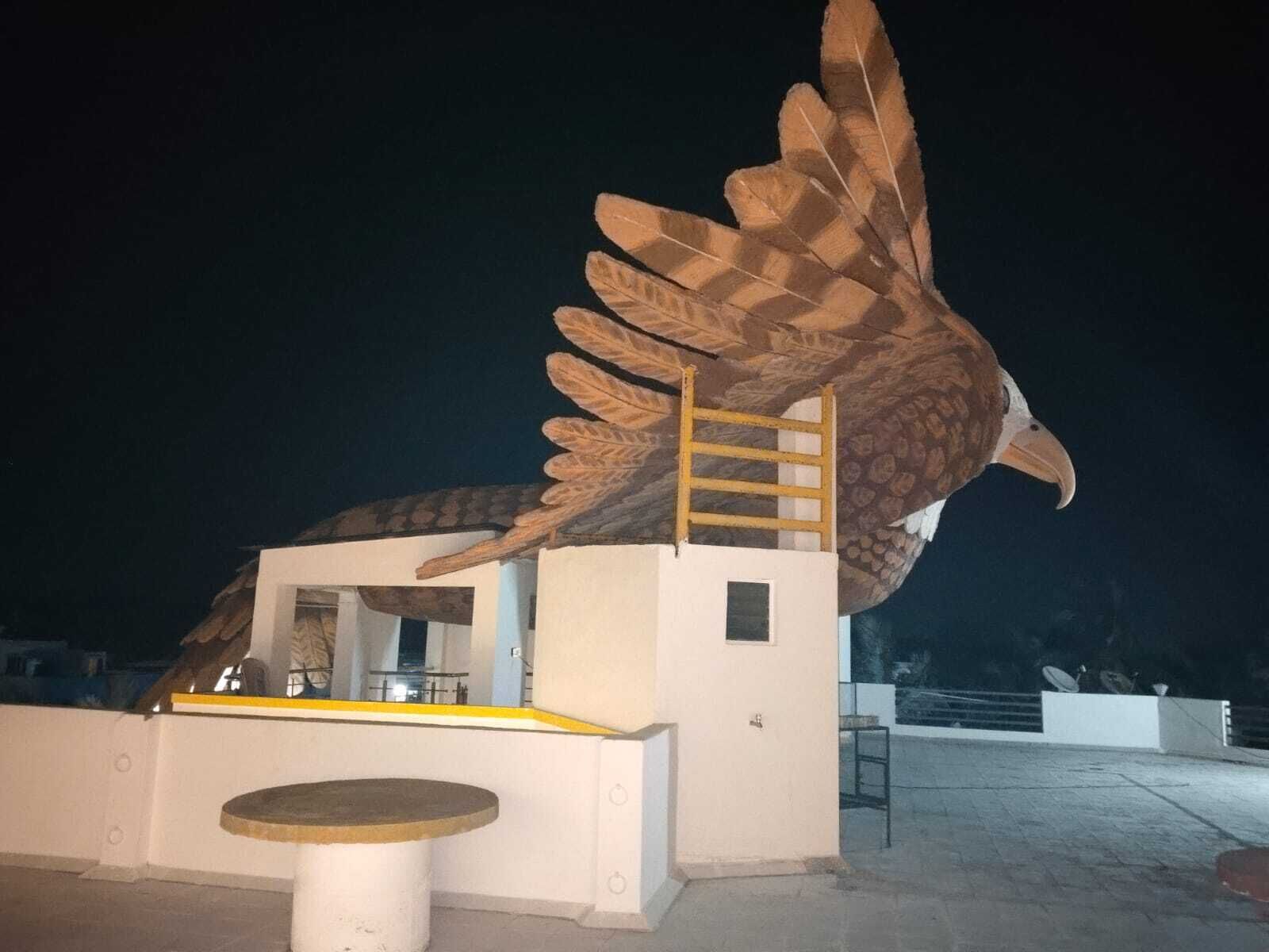 A large bird sculpture with a ladder on its back, a small kiosk, and a table in front.
