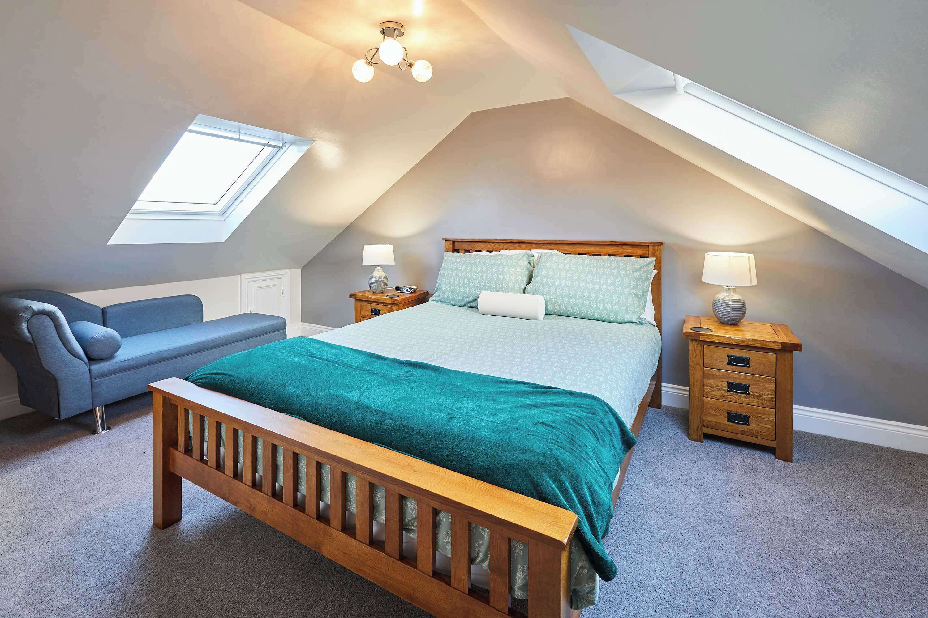 Un dormitorio con techo inclinado, un tragaluz, una cama con colcha verde, un banco de madera, un sofá azul y dos mesitas de noche con lámparas.