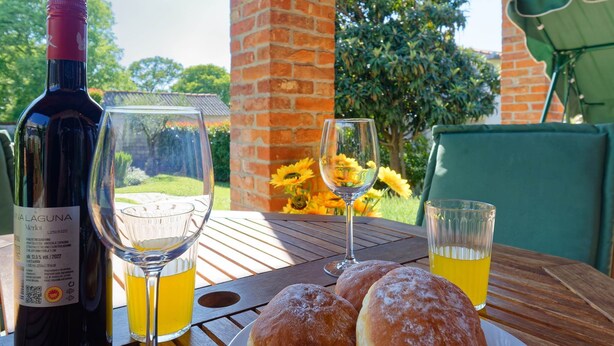 Une bouteille de vin, deux verres à vin, une assiette de pâtisseries et un verre de jus de fruits sur une table en plein air, avec un mur de briques et de la verdure en arrière-plan.