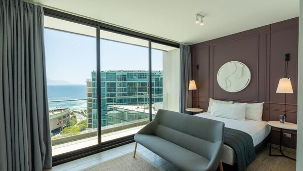 Habitació d'hotel moderna amb una finestra gran, un llit, un sofà i un rellotge de paret rodó.