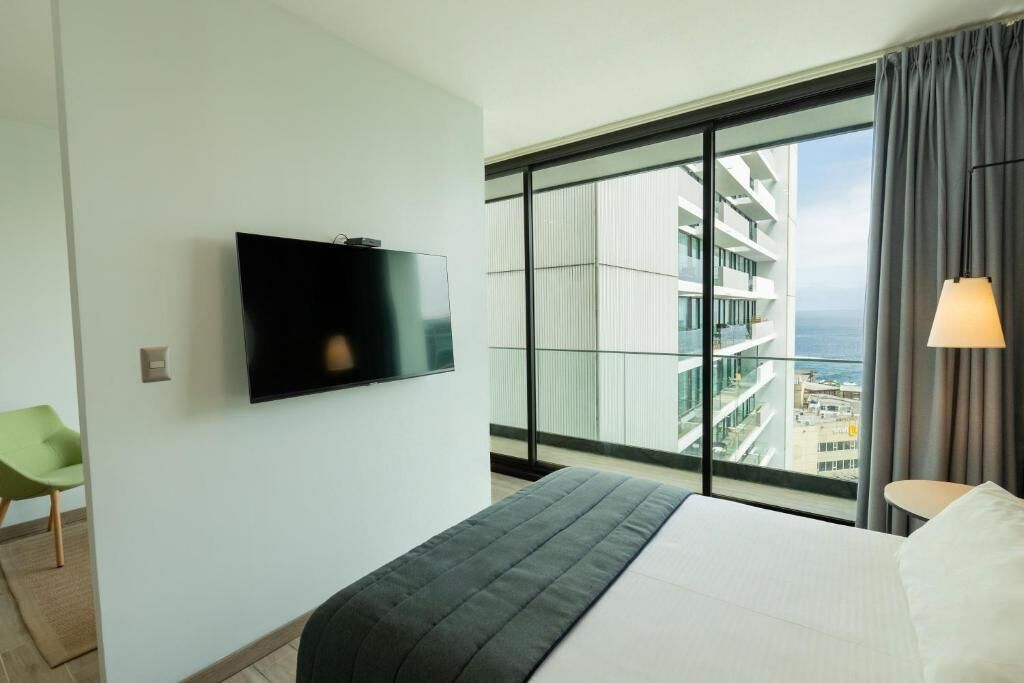 A hotel room with a bed, a TV mounted on the wall, a large window with a view of a building and the sea, and a chair.
