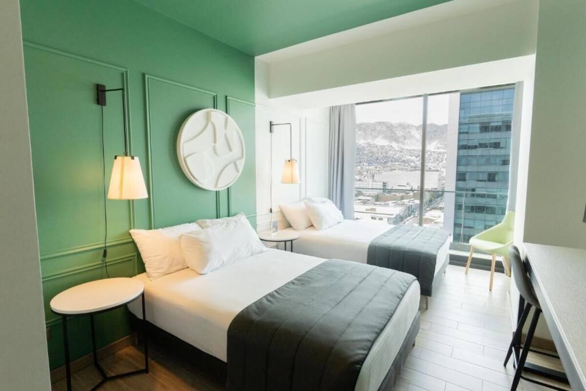 A hotel room with two beds, a round wall clock, a small table, a chair, and a large window offering a city view.