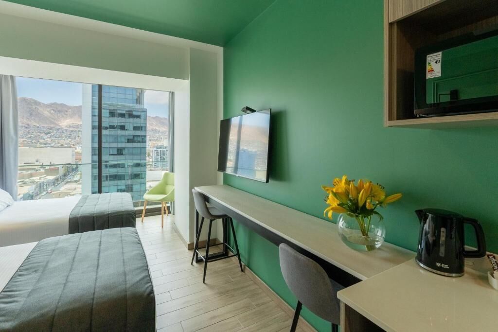 Una habitación de hotel moderna con un ventanal grande que ofrece vistas a la ciudad, un escritorio con silla, una cama y una mesita de noche con un florero.