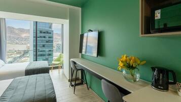 Una habitación de hotel moderna con un ventanal grande que ofrece vistas a la ciudad, un escritorio con silla, una cama y una mesita de noche con un florero.