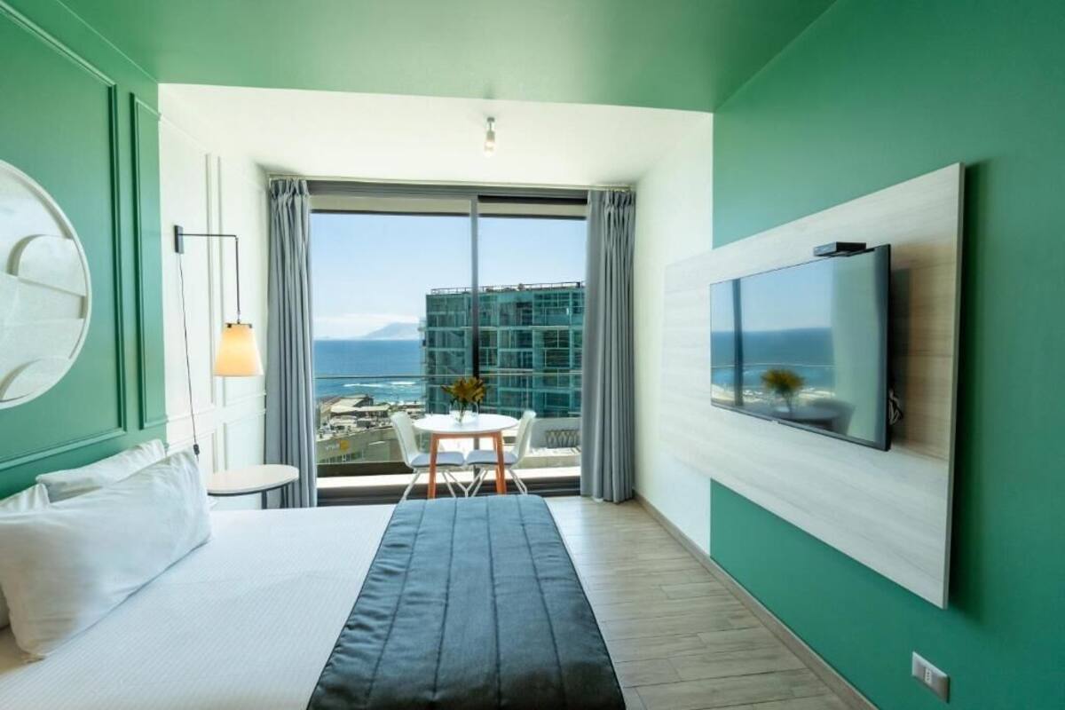 A modern hotel room with a large bed, a TV mounted on the wall, a balcony with a view of the city and ocean, and a small table with chairs.