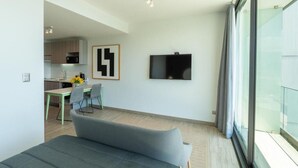 Un saló modern amb un sofà, una zona de menjador amb taula i cadires, una cuina al fons i un televisor fixat a la paret.