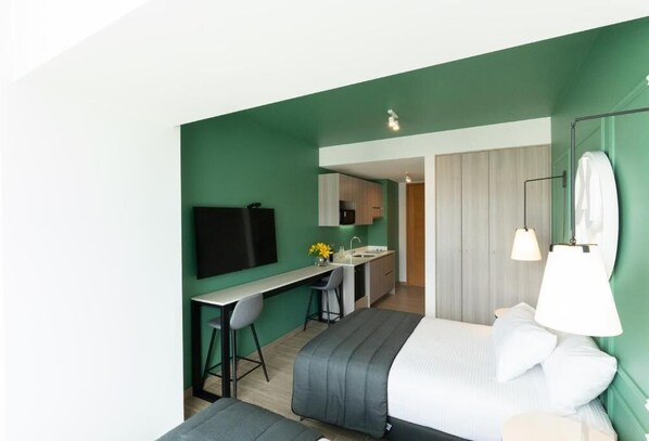 A modern hotel room with a green accent wall, a bed, a desk with a chair, a small kitchenette, and a bathroom with a mirror and lamp. - Studio 56 by Terrado (Antofagasta)
