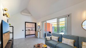 A modern living room with a grey sofa, a wooden coffee table, and a TV on a stand. There is a kitchen in the background with white cabinets and a dining area visible through a glass door.