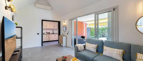 Uma sala de estar moderna com um sofá cinzento, uma mesa de centro de madeira e uma televisão em um móvel. É possível ver uma cozinha ao fundo, com armários brancos e uma área de jantar através de uma porta de vidro.