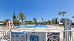 A pool area with a clear sky, palm trees, and a sign indicating COVID-19 norms and a no swimming zone.