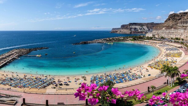 A coastal beach with a curved shoreline, a rocky breakwater, and a building complex on a cliff.