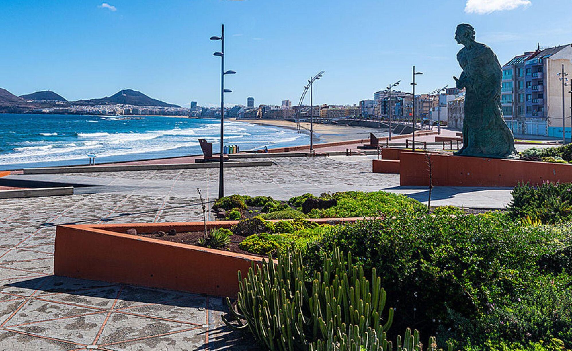 Paseo marítimo con una estatua, cactus y vistas al océano y al paisaje urbano.