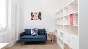 A modern living room with a blue sofa, a white radiator, a framed artwork, a small wooden side table, and a white shelving unit. - Italianway - Cambiasi 8 (Milan)