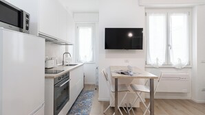 A modern kitchen with white cabinets, a built-in microwave, and a large flat-screen TV mounted above a wooden dining table with two chairs. - Italianway - Cambiasi 8 (Milan)