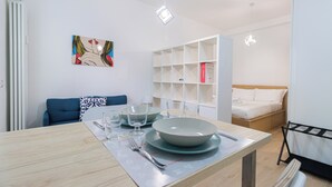 A modern, well-lit room with a dining table set for two, a blue sofa, a bed with white bedding, and a white shelving unit against the wall. - Italianway - Cambiasi 8 (Milan)