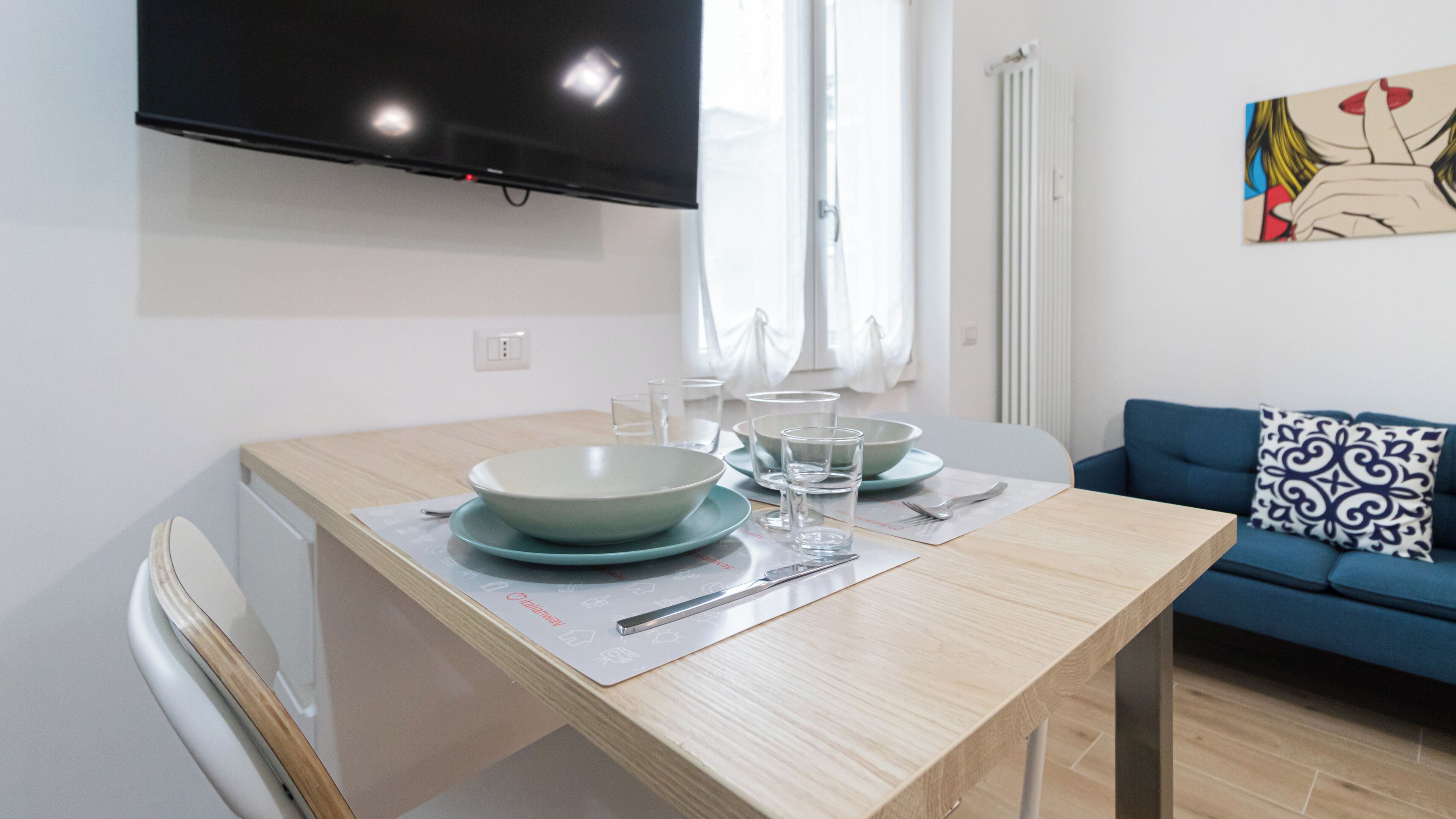 Et spiseområde med et bord dækket til fire personer, et fladskærms-tv på væggen og en blå sofa med en pude med mønster i baggrunden.