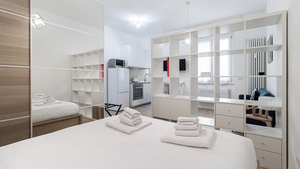A modern hotel room with a large bed, a wardrobe with multiple compartments, a small kitchenette, and a bathroom visible through a glass partition. - Italianway - Cambiasi 8 (Milan)