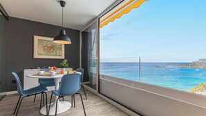 Un comedor moderno con una mesa redonda, sillas azules y una lámpara colgante. Un cuadro enmarcado adorna la pared, y la habitación tiene ventanas amplias con vistas al océano y edificios lejanos.