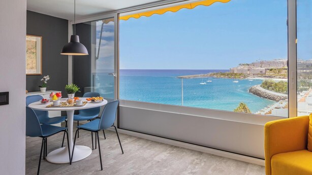 Un comedor moderno con una mesa lista para dos, un ventanal con vista al mar y edificios, y una pared de acento amarilla.