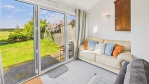 Un salon doté d’une porte-fenêtre coulissante donnant sur un jardin, d’un canapé beige agrémenté de coussins colorés et d’un meuble en bois fixé au mur.