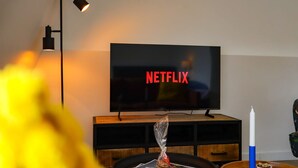 Un salon avec un téléviseur à écran plat affichant le logo de Netflix, un lampadaire noir, un meuble de rangement en bois et une bougie posée sur une table.