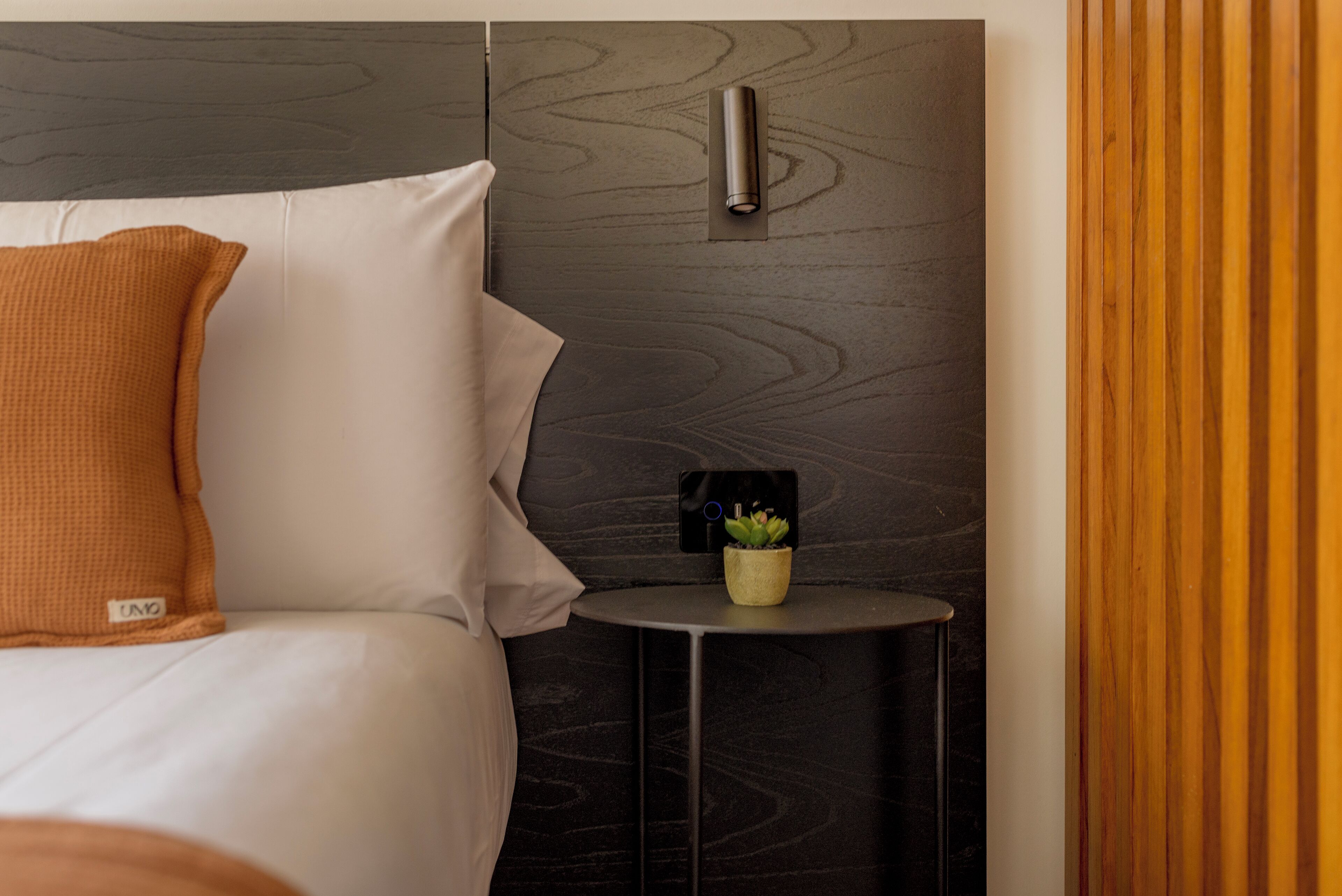 A neatly made bed with a white and orange pillow, a small round table with a plant, and a wall-mounted light fixture.