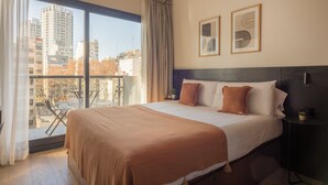 Une chambre d’hôtel avec un grand lit, une vue sur la ville depuis un balcon, et deux œuvres d’art encadrées accrochées au mur.