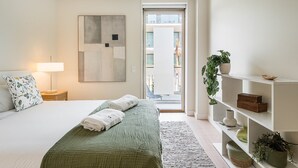 Un dormitorio moderno con una cama, una mesita de noche con lámpara, una colcha verde, un cuadro abstracto de gran tamaño, una planta en maceta y vistas al balcón exterior.
