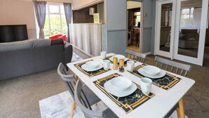 Una sala de estar moderna con un sofá gris, una mesa de comedor con platos blancos y manteles individuales con detalles dorados, y una cocina visible a través de una puerta abierta.