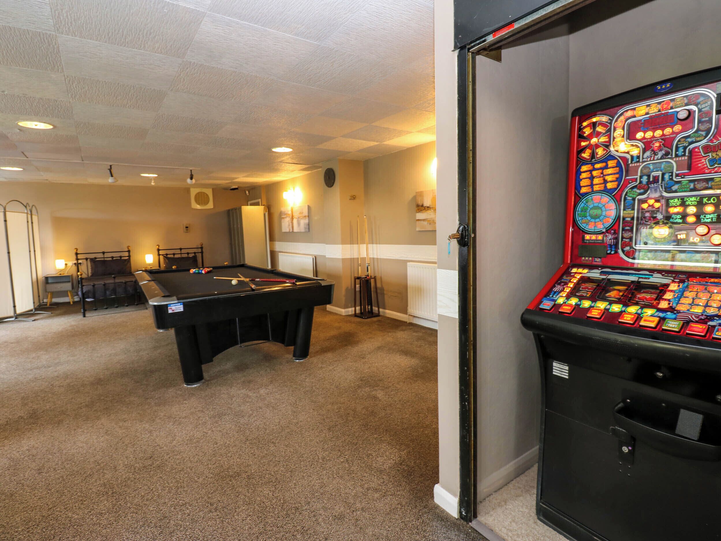 A pool table with billiard balls and cues, a dartboard, and a slot machine in a room with carpeted floors and recessed lighting.