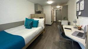 A hotel room with a bed, a desk with a laptop, a chair, and a small table with a lamp and stationery. - Student Only Swanston House (Belfast)
