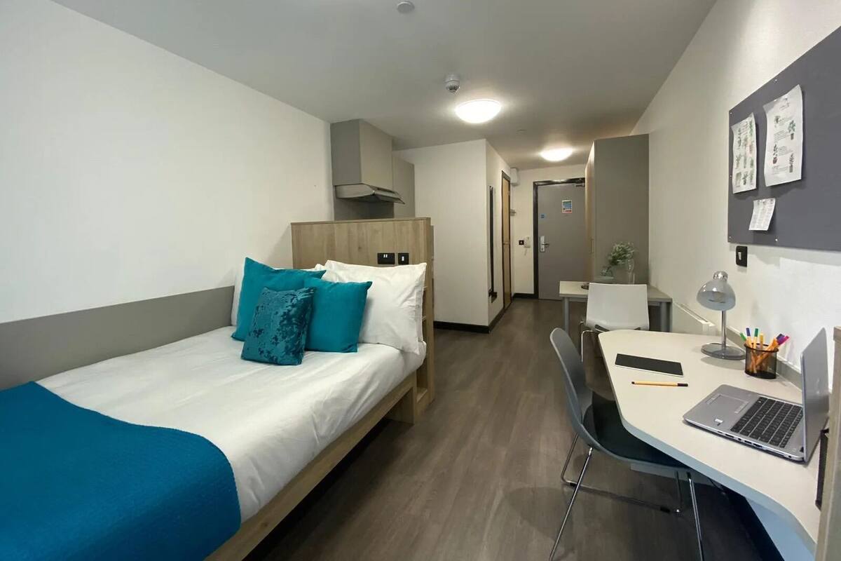 A hotel room with a bed, a desk with a laptop, a chair, and a small table with a lamp and stationery.
