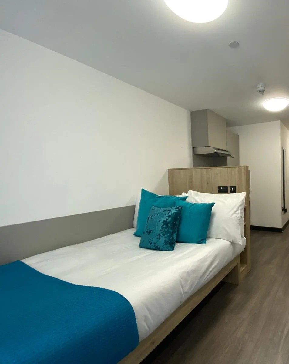 Una cama individual con un edredón azul y almohadas turquesa en una habitación con paredes blancas y suelo de madera.