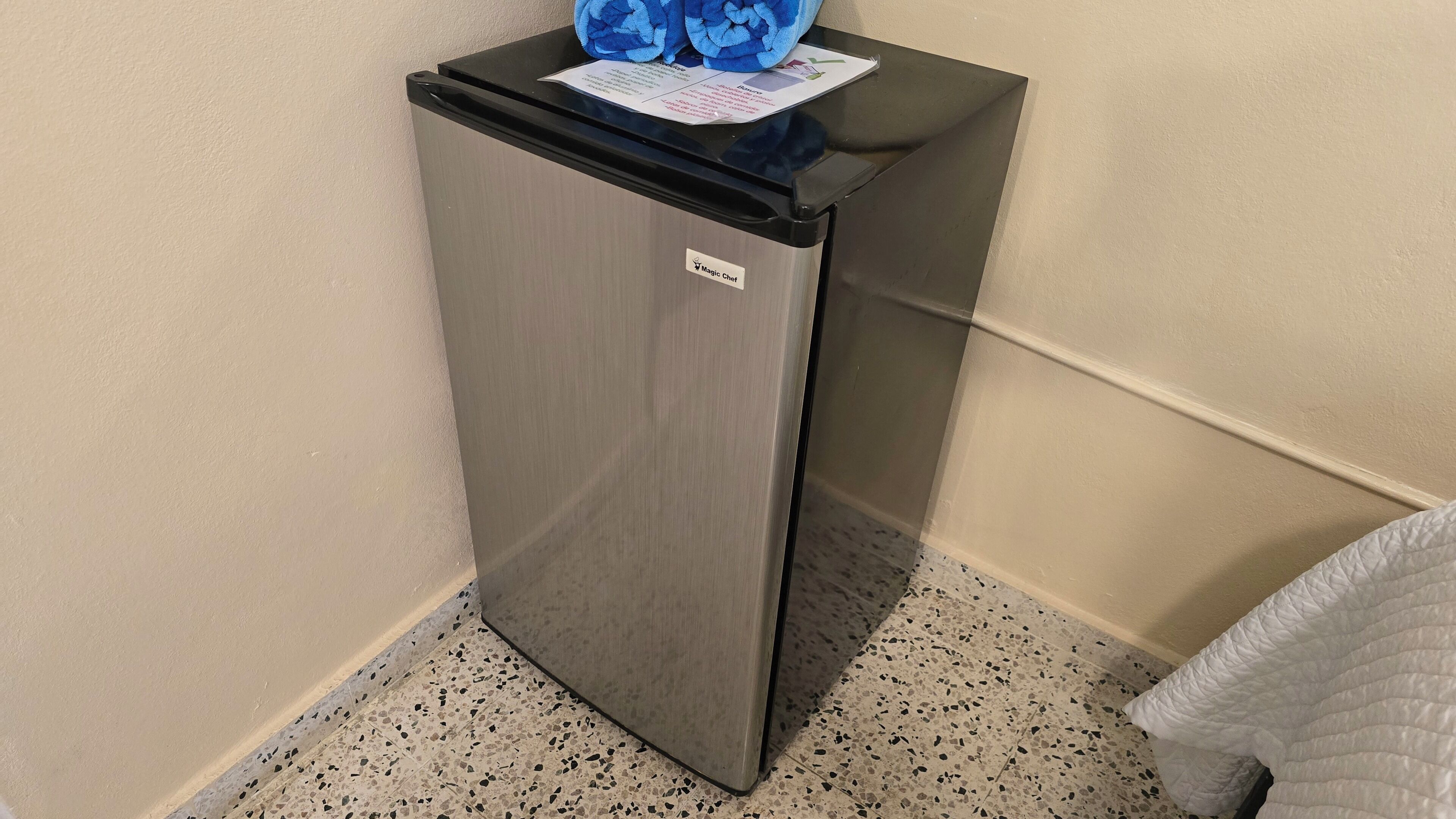 Un petit réfrigérateur, doté d’un dessus noir et d’une porte en acier inoxydable, est placé contre un mur, au-dessus d’un sol à motifs.
