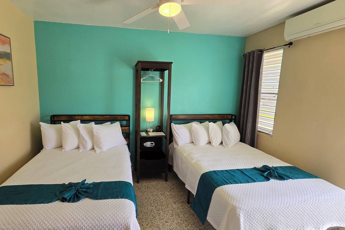 Habitación de hotel con dos camas, paredes verde azulado, ventilador de techo, un cuadro y una ventana con persianas.