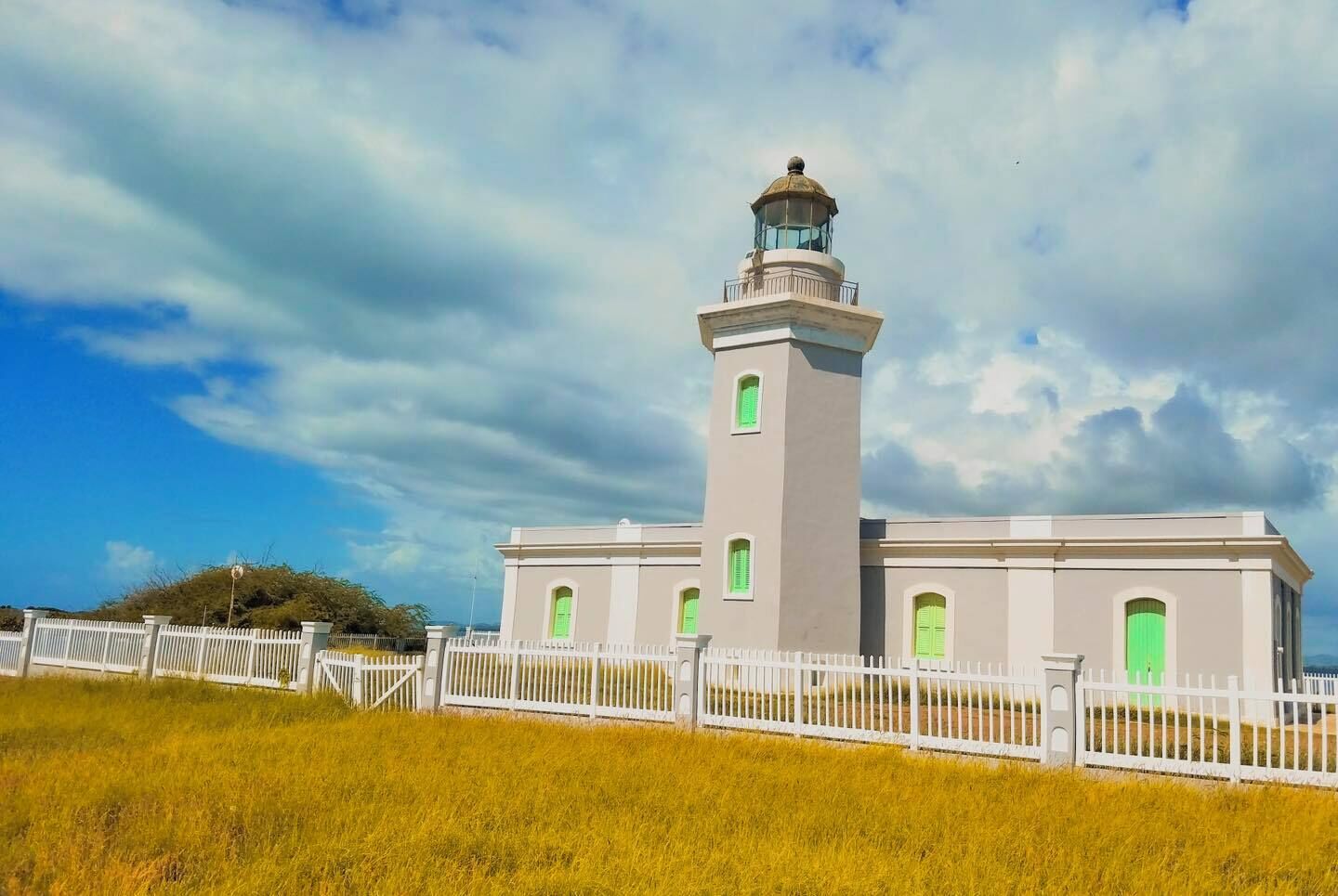 A lighthouse with a white exterior and green shutters, surrounded by a white fence and grassy area.