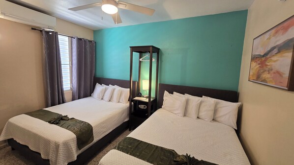 A hotel room with two beds, a nightstand with a lamp, a ceiling fan, and a window with curtains.