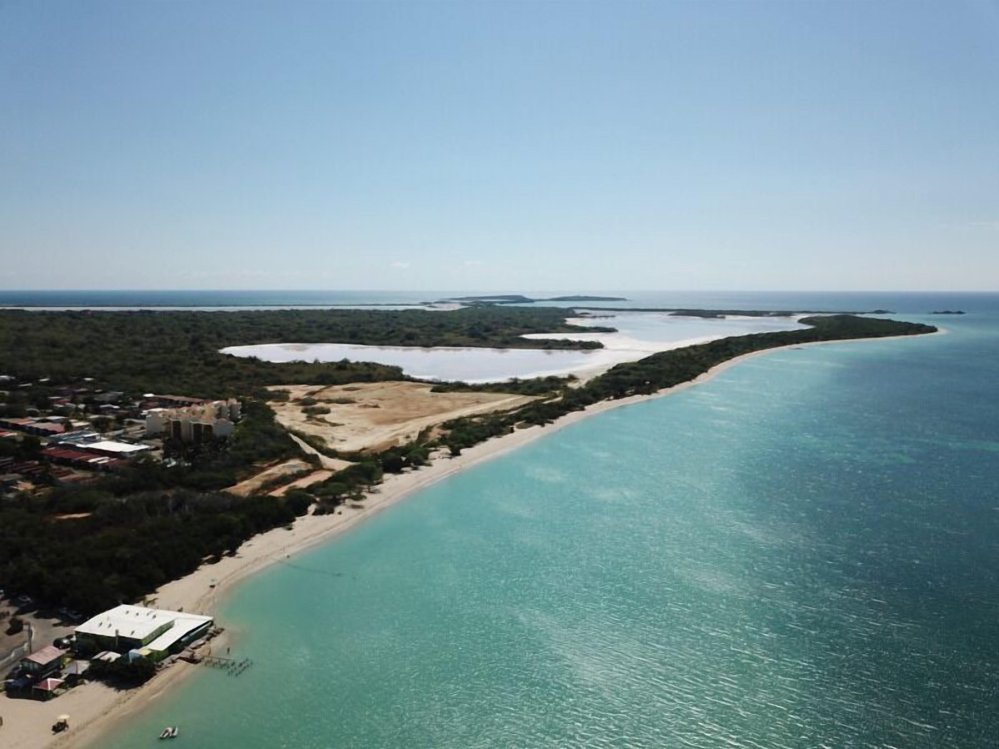 An aerial view of a coastal area with a beach, clear turquoise waters, and a small settlement with buildings and a road.
