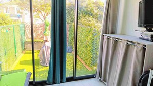 Une pièce dotée d’une grande porte-fenêtre coulissante donnant sur un jardin. On y trouve un lit avec un oreiller vert, un téléviseur posé sur un meuble et une petite table avec des livres et une télécommande.