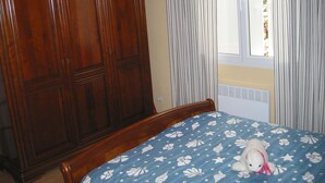 Un dormitori amb un armari de fusta gran, un llit amb edredó floral blau i una finestra amb cortines.