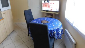 Una zona de menjador petita amb una taula rodona coberta per un mantell blau, dues cadires negres, un forn blanc i una televisió sobre un moble.