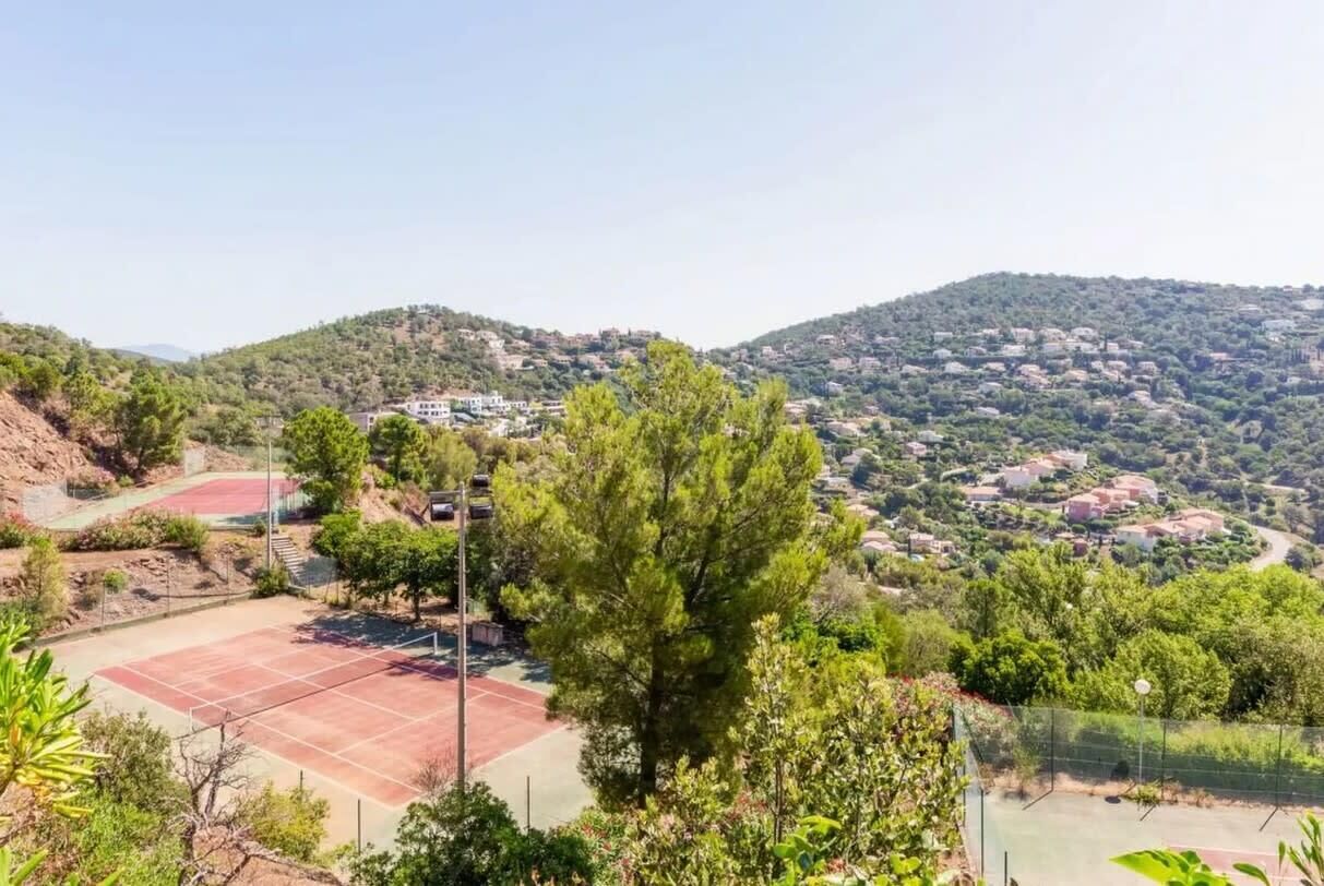 A tennis court surrounded by greenery and hills with buildings in the background.
