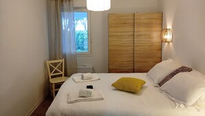 A bedroom with a bed, a chair, a window with curtains, a wall-mounted heater, and a bedside lamp.