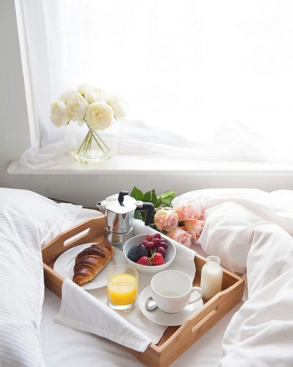 A breakfast tray with croissant, fruit, coffee, and juice on a bed with white bedding and a window with white flowers.