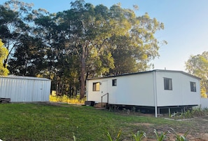 A mobile home with a small porch and a shed in a grassy area with trees.