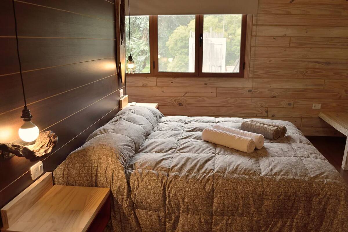 A bedroom with a bed, a nightstand, a wooden bench, and a window with a view of trees.