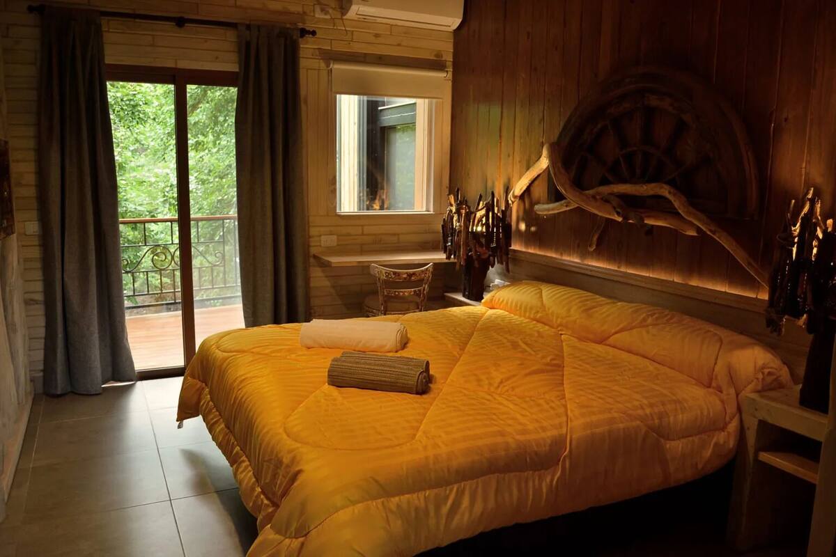 A large bed with a yellow bedspread, a wooden headboard with a wheel, a small table, and a chair.