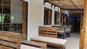 Ein überdachter Außenbereich mit Holzbänken und -tischen, einer Steinwand und einer Bambuskonstruktion.